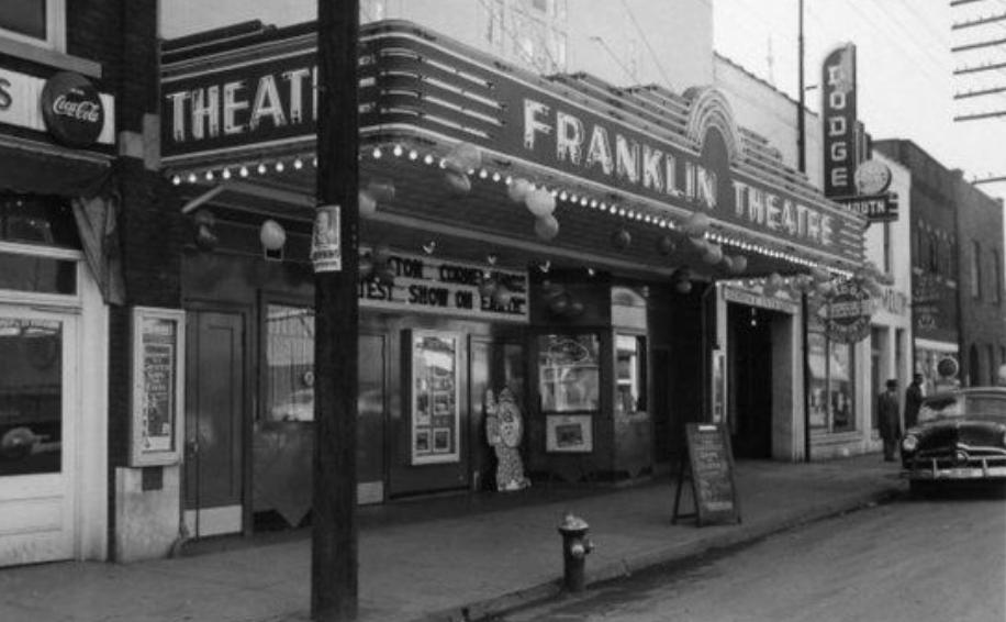 Historic black-and-white photo of the Franklin Theatre marquee on Main Street, decorated with balloons, with a vintage car parked out front