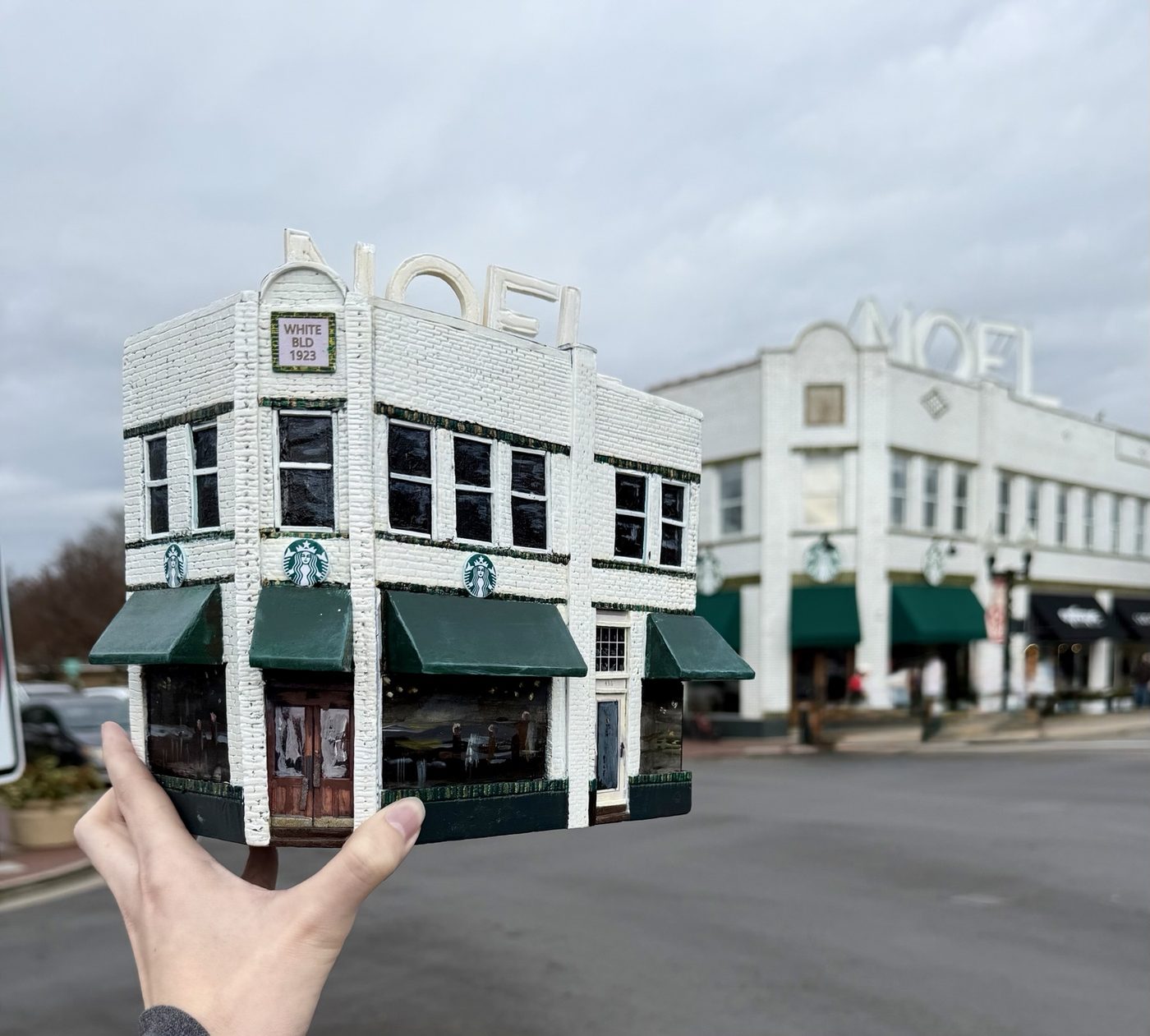 A hand-painted miniature of the White Building held up in front of the real building in downtown Franklin, TN